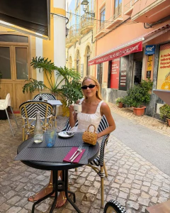 Mulher sentada em mesa de café ao ar livre, vestindo vestido branco curto com alças e usando óculos escuros