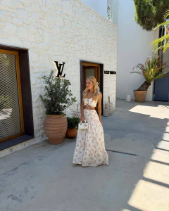 Mulher loira usando conjunto Fiorenza branco com estampa floral e bordados, composto por top cropped com detalhe em guipir e saia longa plissada, em frente à parede branca com logo Louis Vuitton