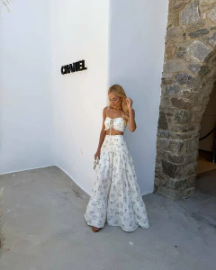 Mulher loira usando conjunto branco estampado com top de alças finas e saia longa plissada, em ambiente externo com parede branca e arco de pedra