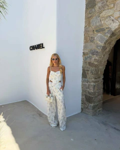 Mulher loira usando conjunto branco estampado Yasmin, formado por top estilo corset e calça larga, posando ao lado de parede branca com logo Chanel