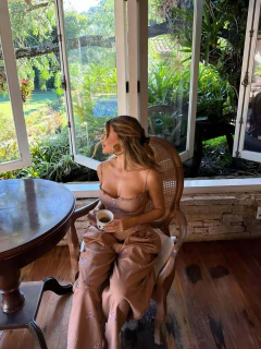 Mulher sentada em cadeira de madeira perto de mesa redonda, vestindo conjunto floral de linho em tom bege com estampa delicada, segurando uma xícara de café, em ambiente interno com grandes janelas abertas e vista para jardim verde.