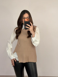 Mulher vestindo conjunto camisa tricot bege com camisa branca por baixo e calça preta, tirando selfie com celular