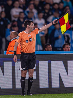 Árbitro de fútbol en cancha señalando con bandera de asistente, vestido con camiseta naranja y short negro Skechers