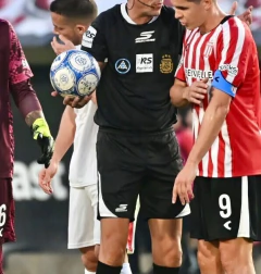 Árbitro de fútbol vistiendo uniforme negro Skechers con short y camiseta, sosteniendo un balón oficial en una cancha durante un partido