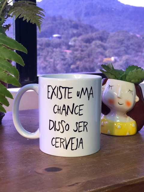 Caneca branca com a frase "EXISTE uma CHANCE DISSO SER CERVEJA" em destaque, ao lado de um vaso decorativo.